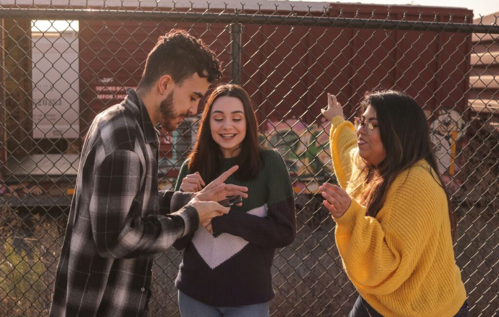 A guy and two girls using a phone by the wire fence of a trainyard